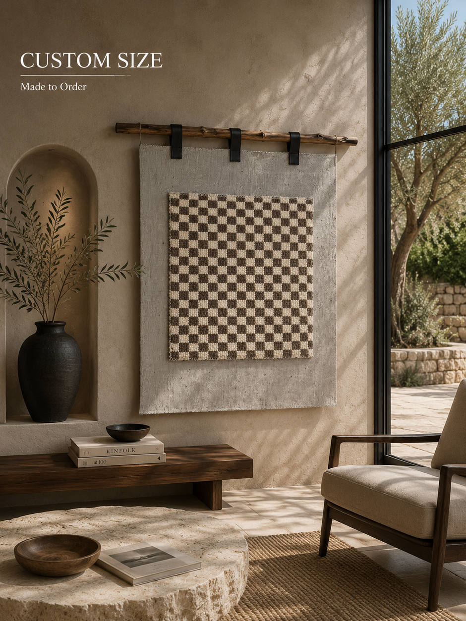 A custom size wall hanging featuring a black and beige checkerboard pattern on a textured gray background, displayed in a modern, sunlit room with a chair, wooden bench, and large window overlooking a garden.