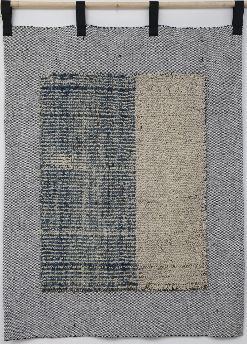 A square woven textile wall hanging or rug in gray, with a central rectangular panel split vertically into a blue-and-white textured left side and a beige right side, suspended from a wooden rod with black clips against a white wall.