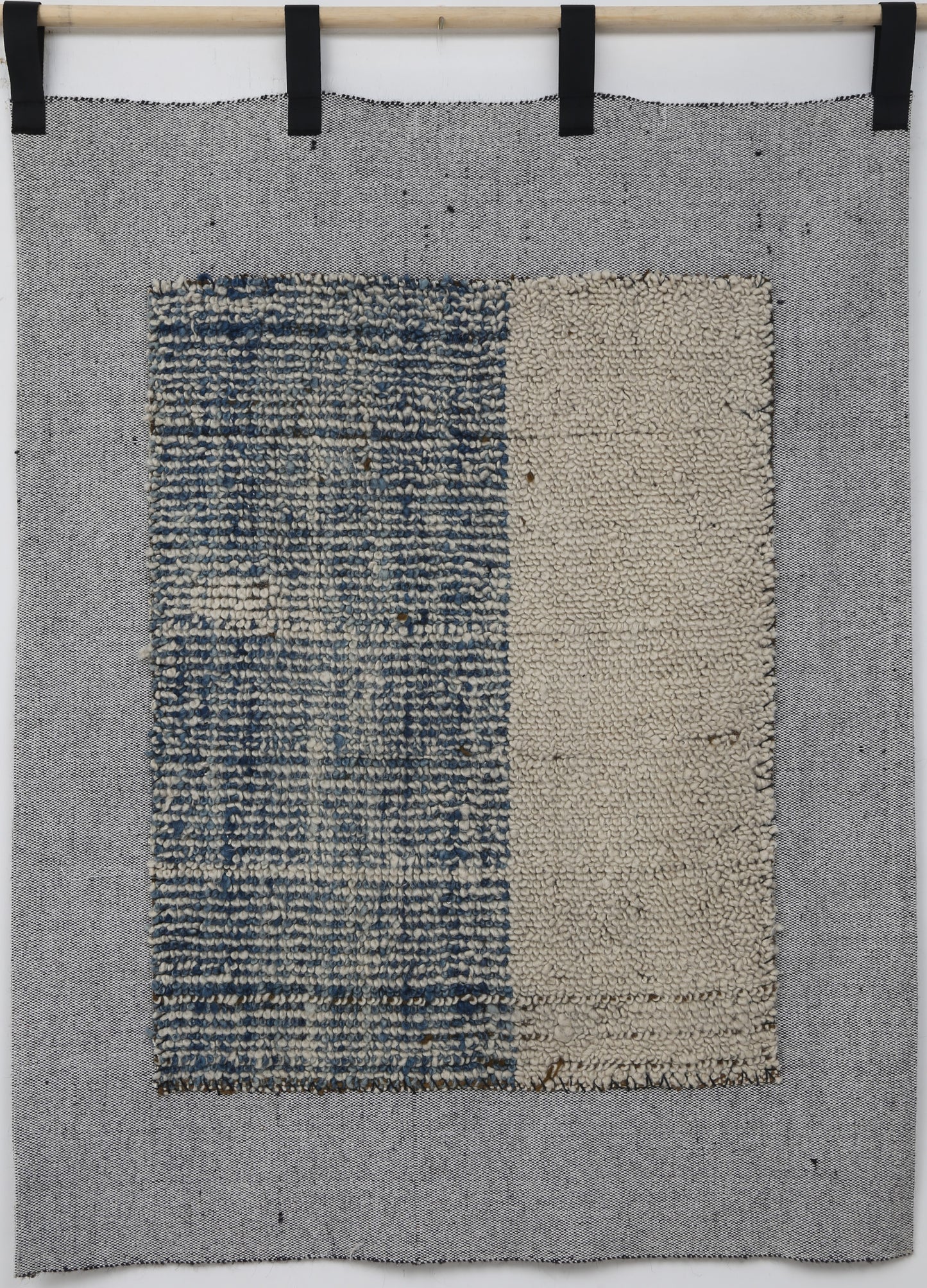 A square woven textile wall hanging or rug in gray, with a central rectangular panel split vertically into a blue-and-white textured left side and a beige right side, suspended from a wooden rod with black clips against a white wall.