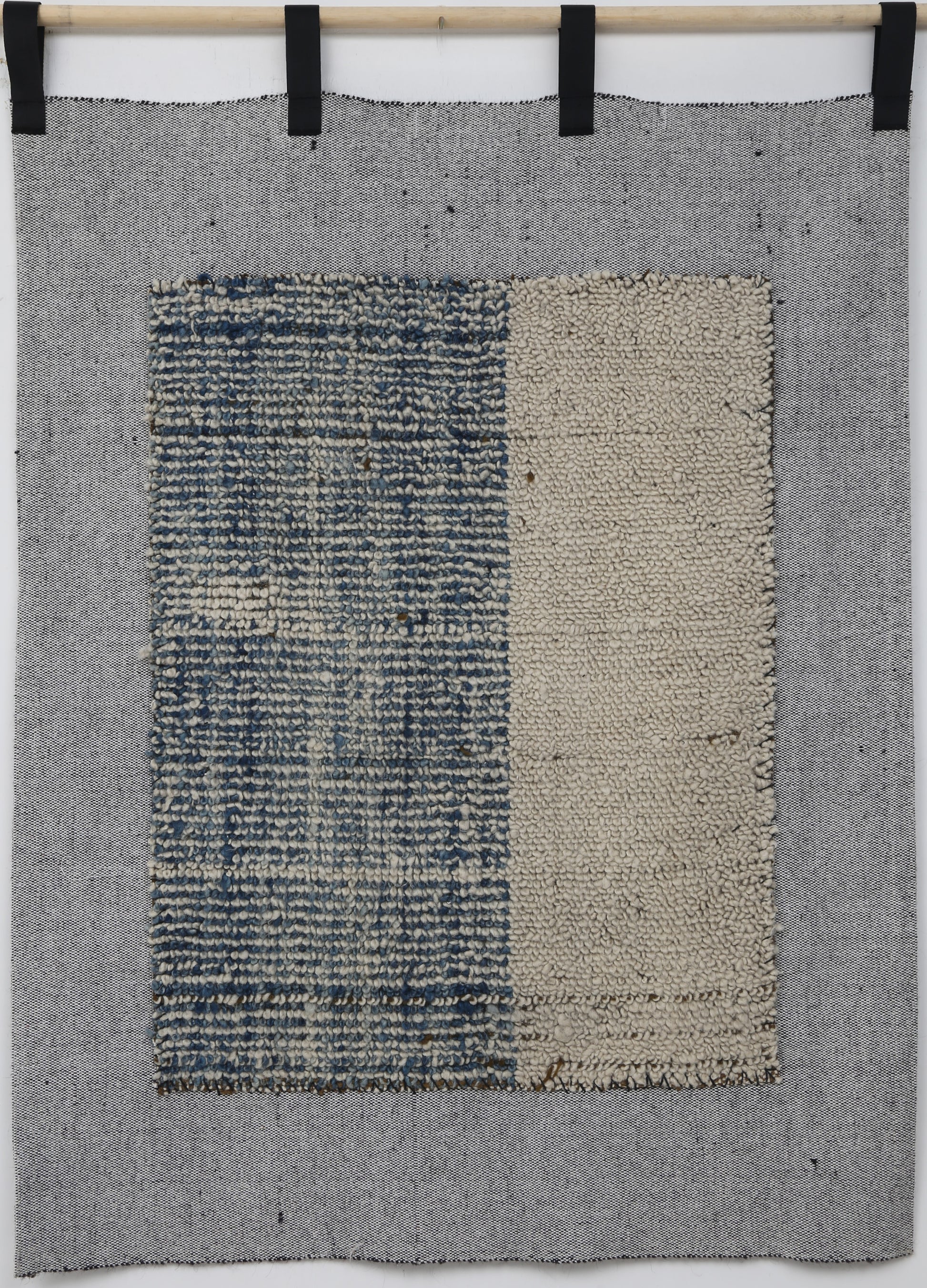 A square woven textile wall hanging or rug in gray, with a central rectangular panel split vertically into a blue-and-white textured left side and a beige right side, suspended from a wooden rod with black clips against a white wall.