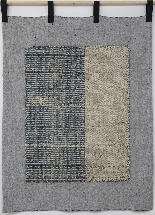 A square woven textile wall hanging or rug in gray, with a central rectangular panel split vertically into a blue-and-white textured left side and a beige right side, suspended from a wooden rod with black clips against a white wall.