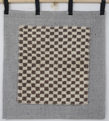 A square woven textile panel with a brown and cream checkerboard pattern, mounted on a larger gray fabric backdrop with black hanging tabs at the top.
