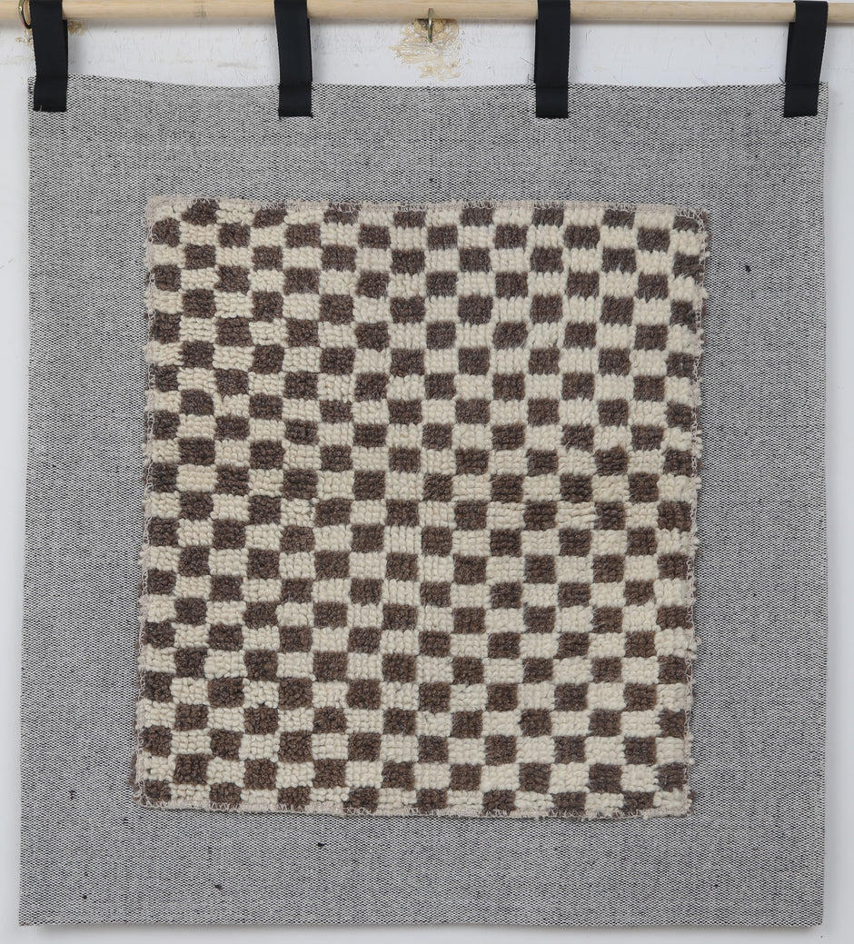 A square woven textile panel with a brown and cream checkerboard pattern, mounted on a larger gray fabric backdrop with black hanging tabs at the top.